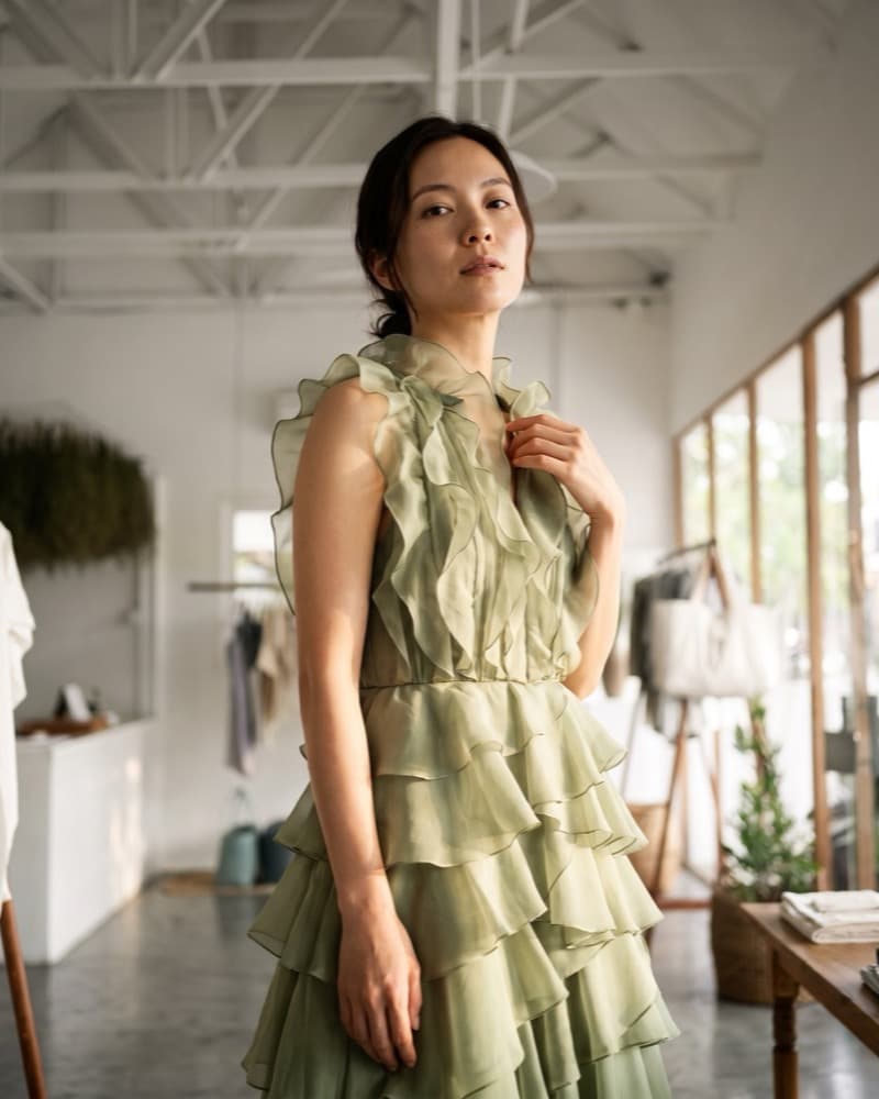 A sage tiered organza gown worn in the boutique, the model's hand at the ruffled neckline, clothing rails soft behind.