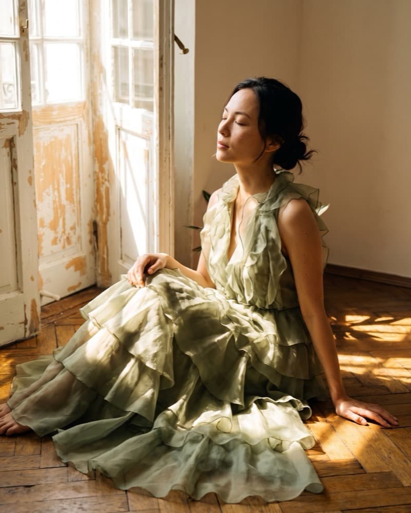 The same gown on the same model in a different room: seated on parquet by a peeling casement door in low golden light.